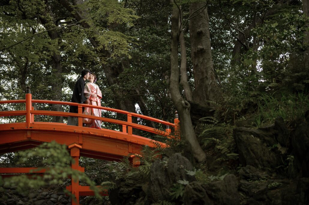 On an arched vermilion bridge in a deep green garden, a groom in montsuki kisses the cheek of his bride in a pink iro-uchikake.