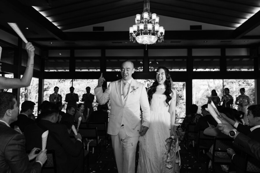 The bride and groom walk back up the aisle as newlyweds under a chandelier, guests raising their arms, in black and white.