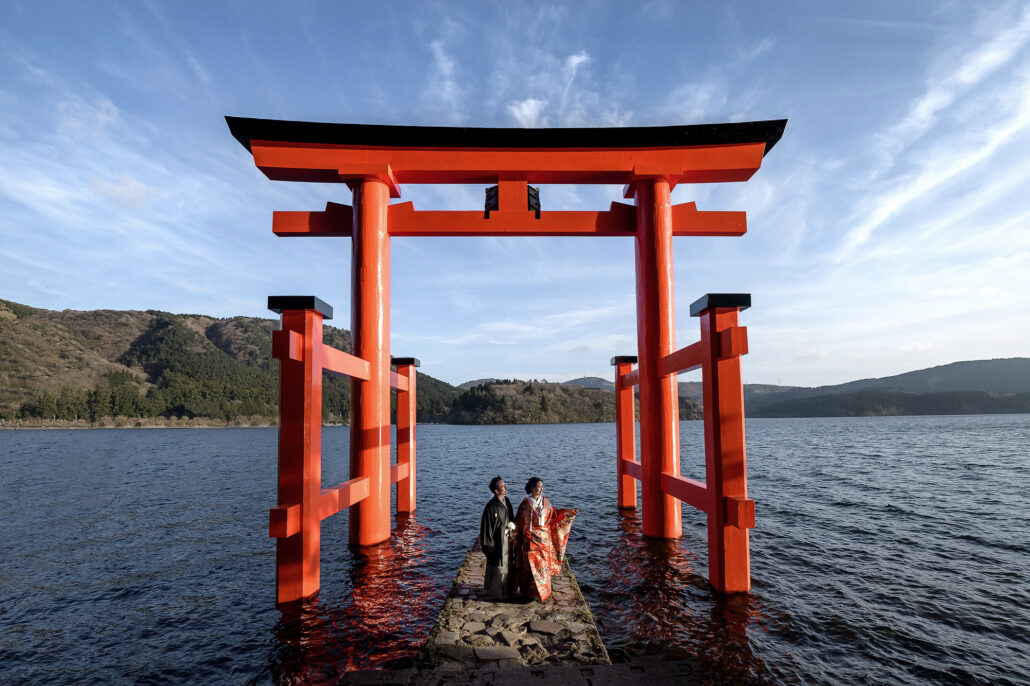 Under the great vermilion torii standing in Lake Ashi, Hakone, a bride in a red iro-uchikake and her groom in montsuki look out over the water.