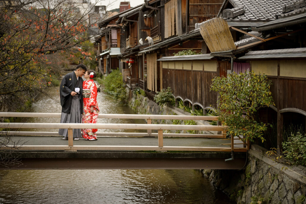 On a small bridge over the Shirakawa canal in Gion, Kyoto, a bride in a red furisode and her groom in montsuki and hakama touch foreheads.