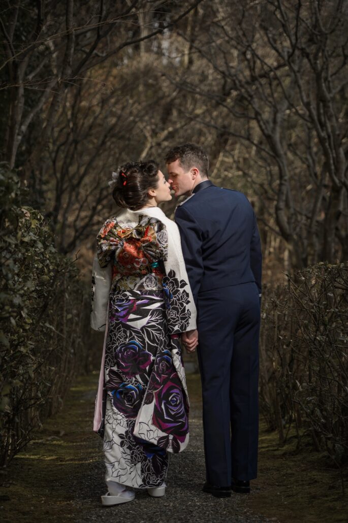 On a hedged garden path, a bride in a white furisode printed with black and purple roses, a gold obi bow at her back, kisses her groom.