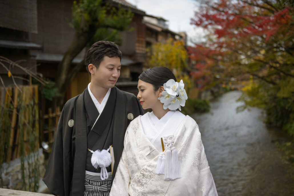 By the Shirakawa in Gion, Kyoto, a bride in a white shiromuku with white orchids in her hair smiles at her groom in black montsuki, autumn colour behind.