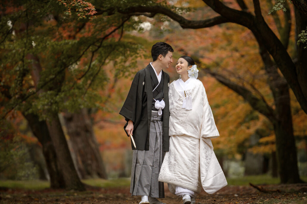 Laughing together, a bride in a white shiromuku and her groom in montsuki and hakama walk an avenue of autumn maples in Kyoto.