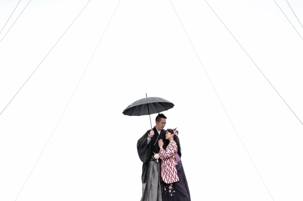 Under a black umbrella in the rain, a bride in a yagasuri arrow-feather kimono and purple hakama rests against her groom in montsuki.