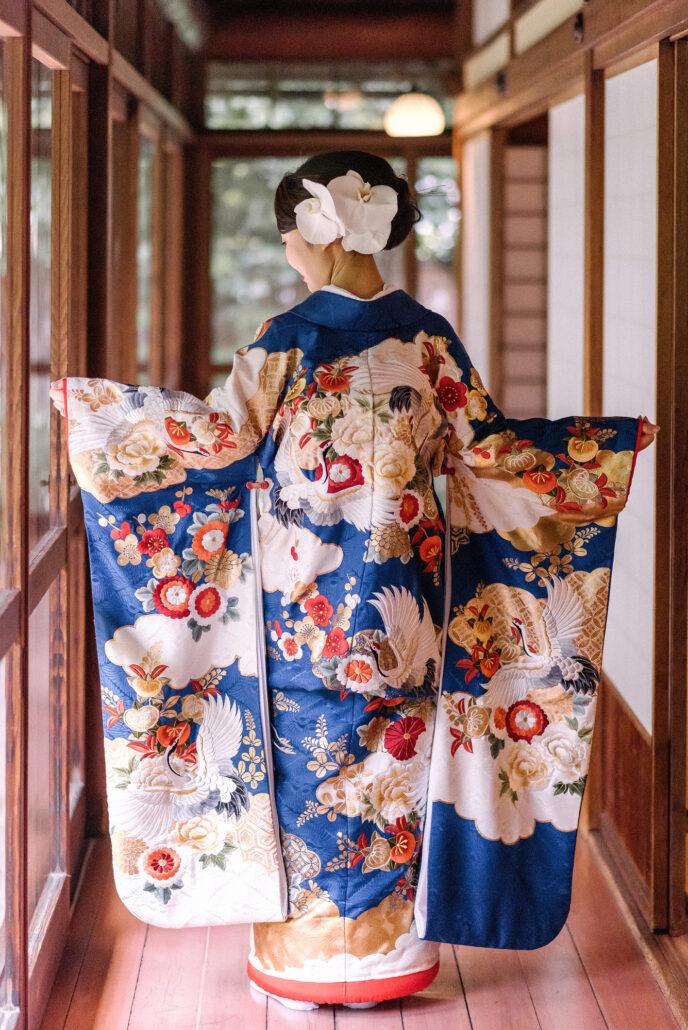 Arms held wide in a wooden corridor, a bride shows the back of her blue and ivory iro-uchikake, cranes and peonies trailing to a padded red hem.