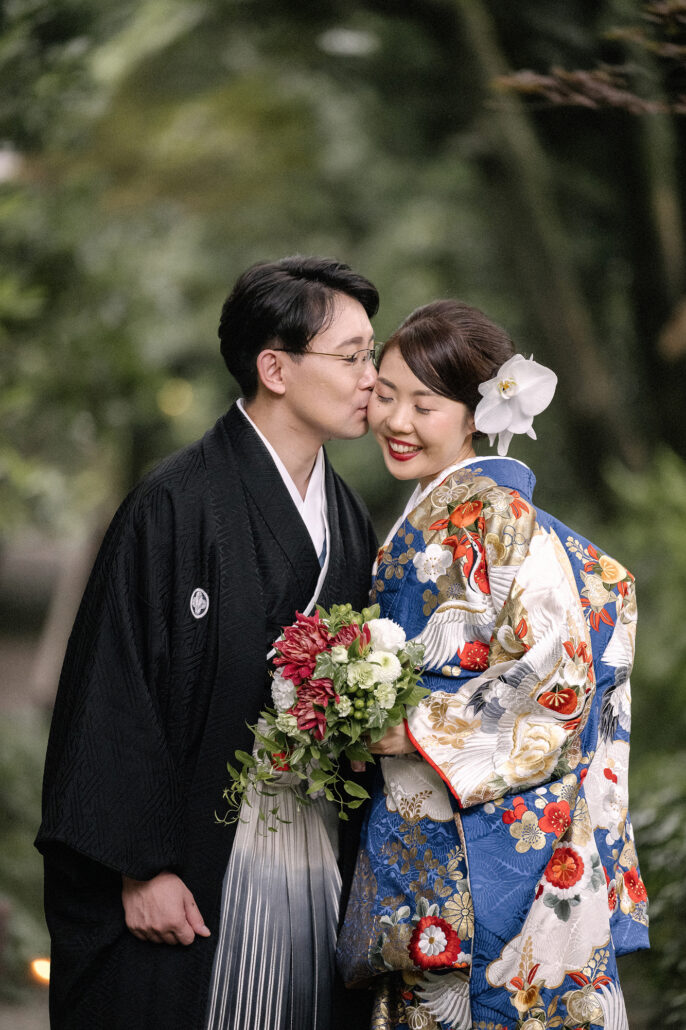 In a green garden, a groom in black montsuki kisses the hair of his bride in a blue and ivory crane iro-uchikake, a red and white bouquet in her hands.