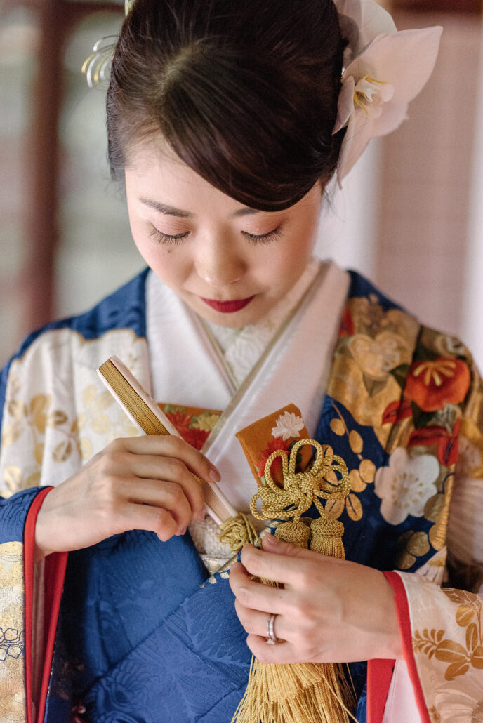Close in, a bride adjusts the gold cords and hakoseko case at the collar of her blue crane iro-uchikake, a white orchid in her hair.