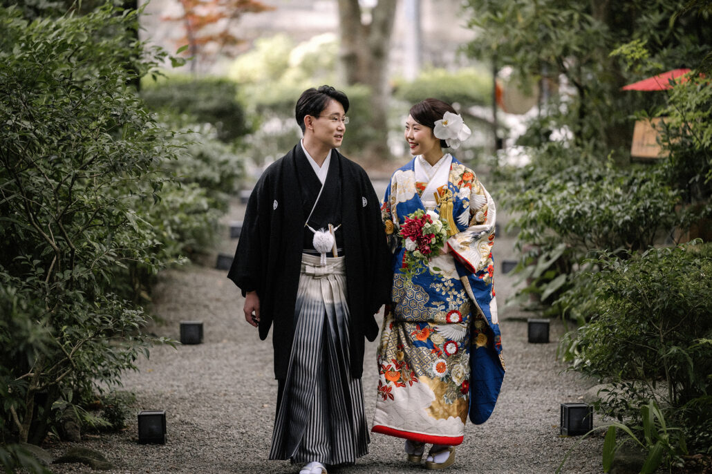 A bride in a blue and ivory crane iro-uchikake and her groom in montsuki and hakama walk a garden path, red parasols behind them.