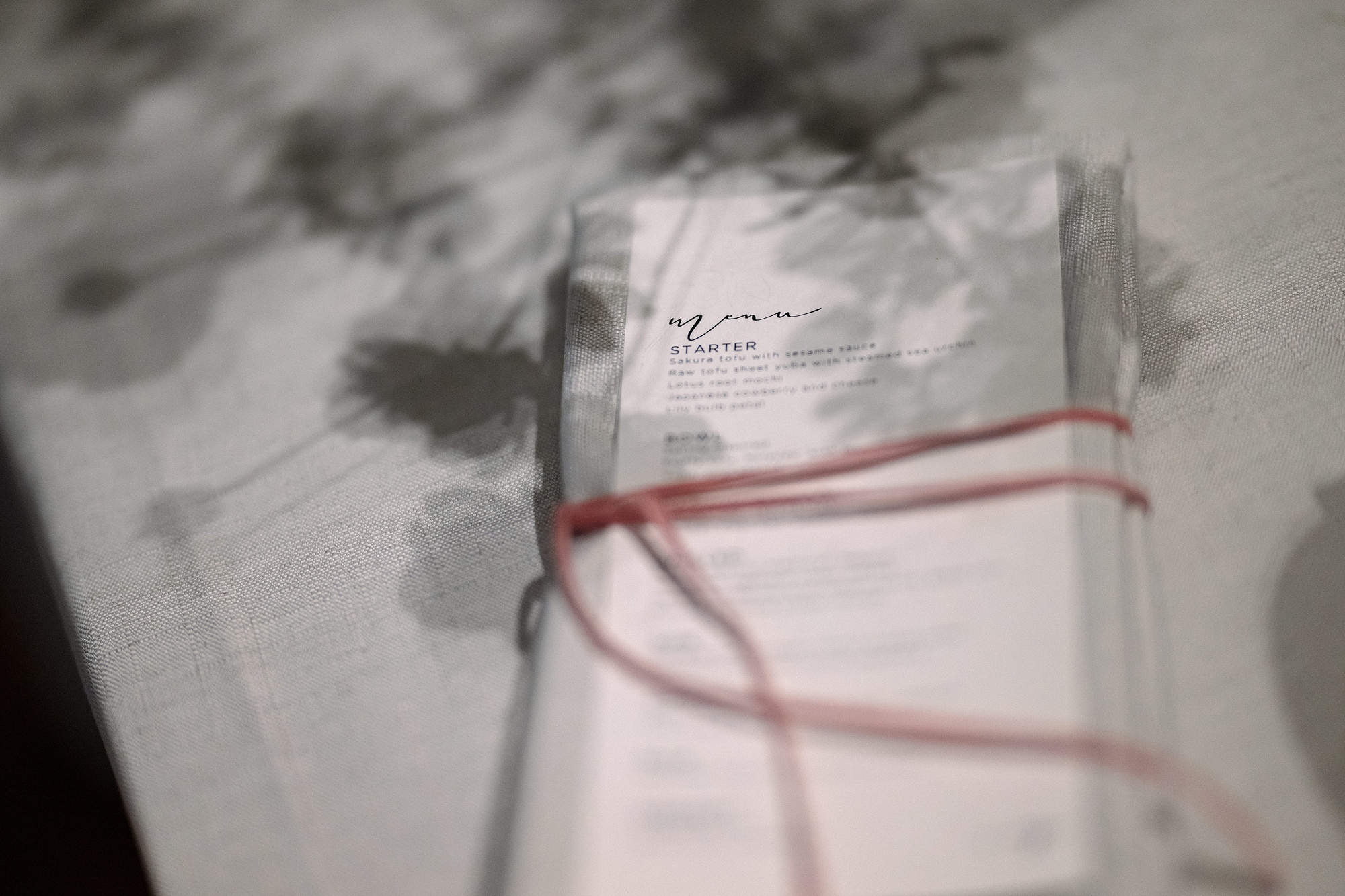 A wedding menu card tied with red cord rests on linen as the shadow of cherry blossom falls across it, at a ryokan wedding in Japan.