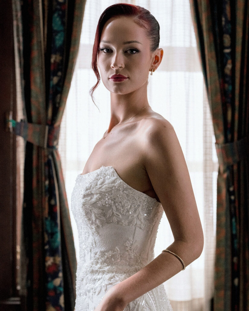 The bride in a strapless lace gown looks into the camera in soft window light between patterned curtains, ready for her destination wedding in Japan.