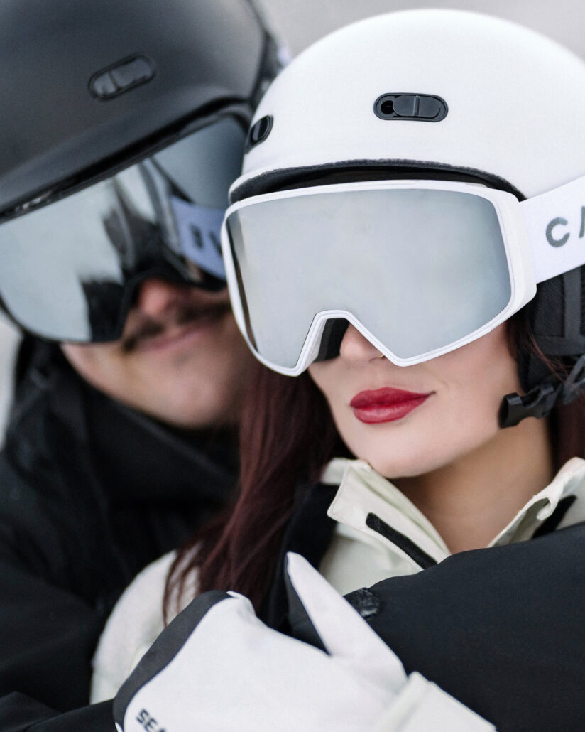 A close portrait of the bride in a white ski helmet and mirrored goggles with red lipstick, the groom beside her at the post wedding shoot.