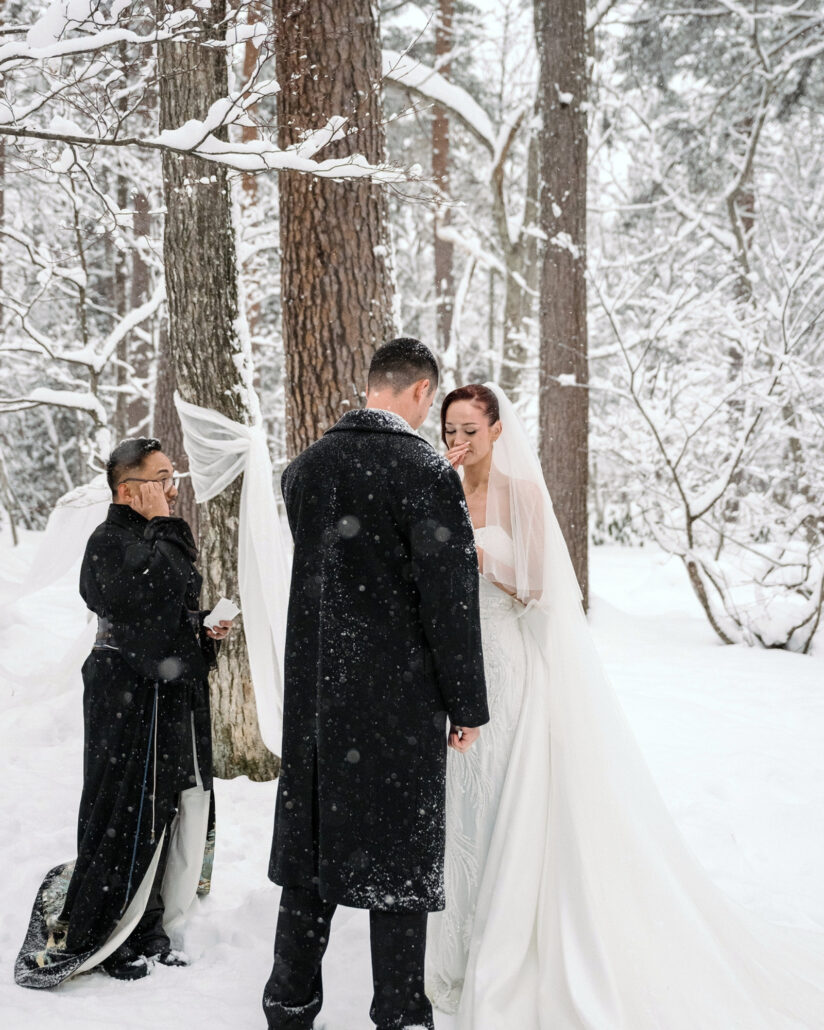 The officiant in a black kimono style robe wipes his eyes as the bride lifts a hand to her face before the groom at their forest wedding in Hakuba.