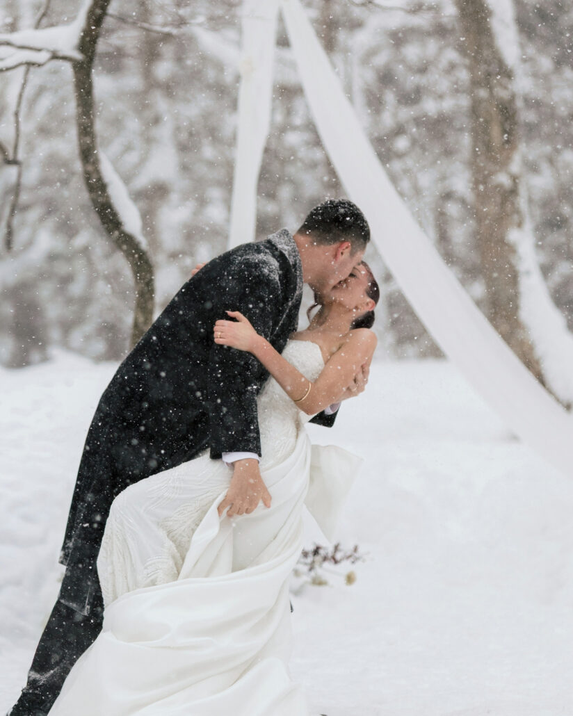 The groom dips the bride for a kiss in the falling snow, white ribbons hanging from the trees of the forest ceremony.