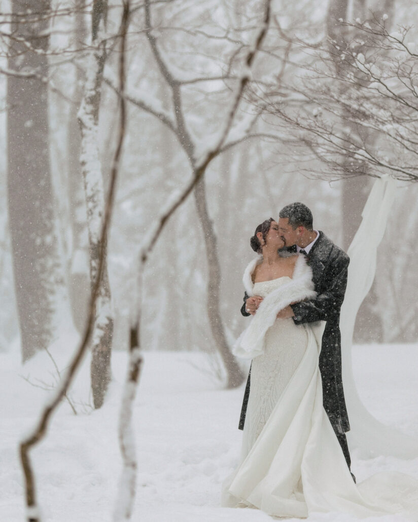 The groom in a dark overcoat kisses the bride in her white stole among snow covered birch trees at their winter wedding in Japan.