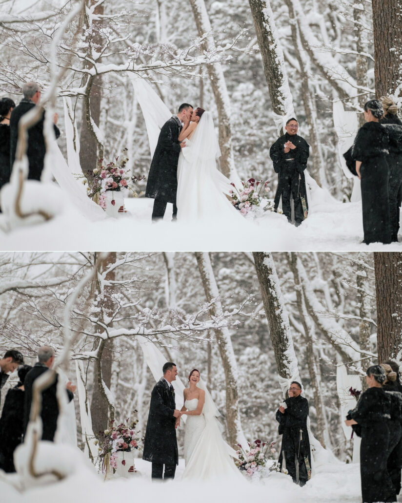The couple kiss before the officiant at their forest ceremony in falling snow, guests in long black coats standing among the trees.