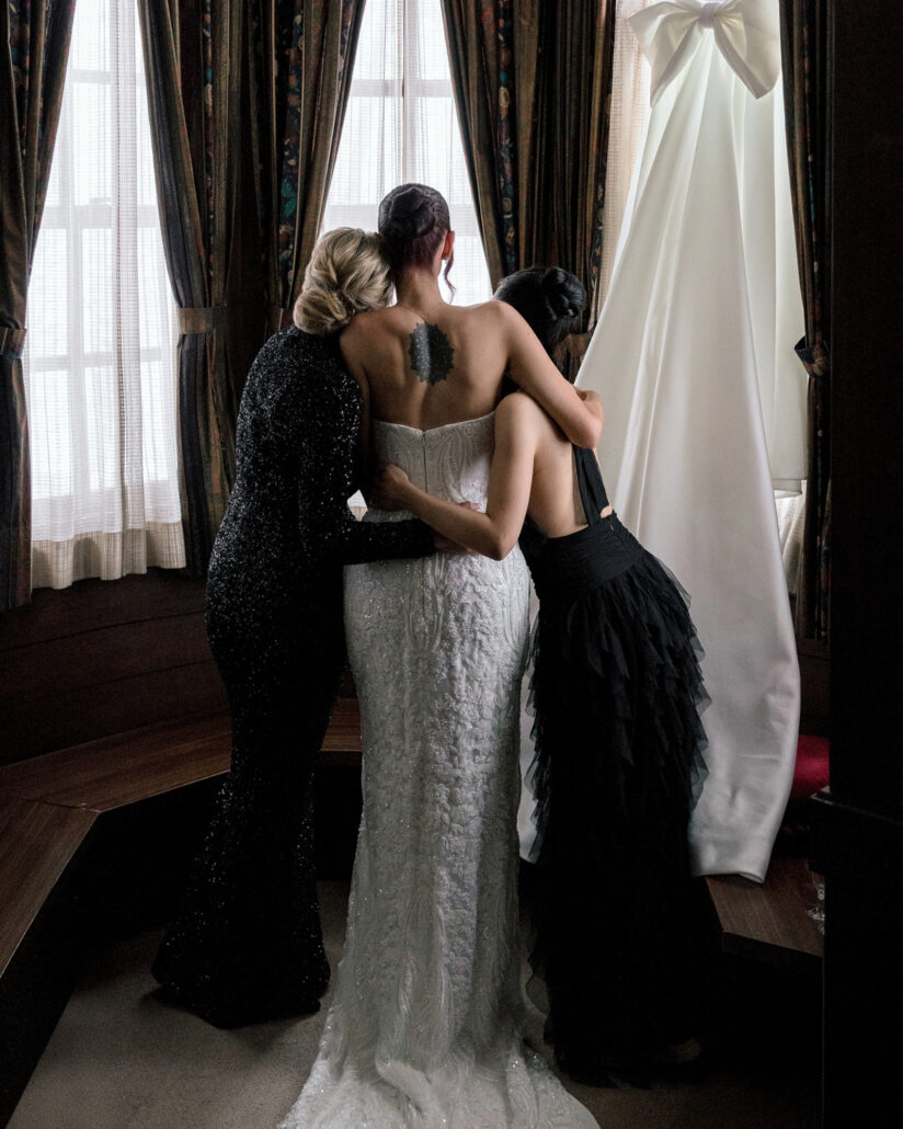 The bride in her lace dress hugs two friends by the chalet window, her wedding gown hanging behind them.