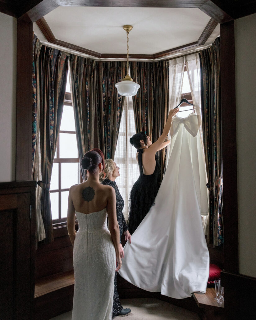 The bride in a fitted lace dress watches as a friend lifts her wedding gown down from where it hangs in the bay window.