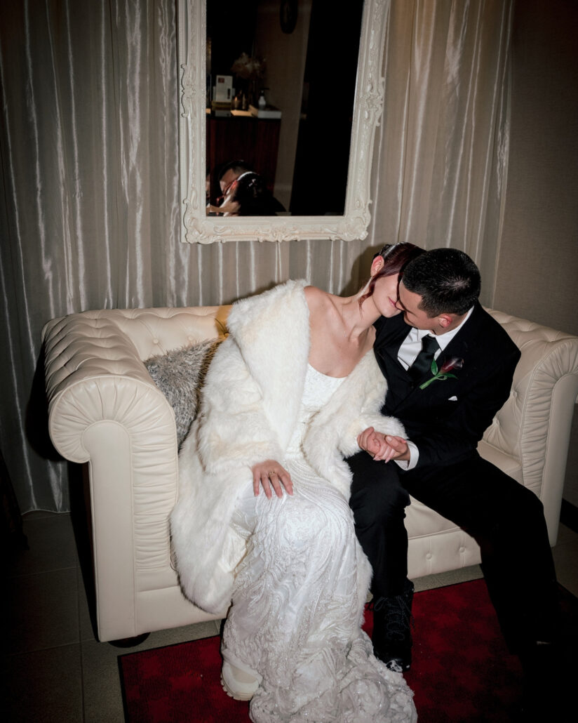 The groom leans in close to the bride in her white fur coat on a white chesterfield sofa beneath an ornate mirror after their snow wedding in Japan.