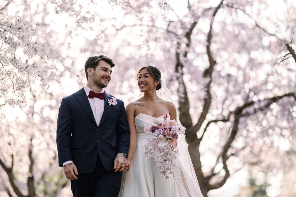The bride and groom walk hand in hand beneath white cherry blossom in an Aomori grove, her bouquet in soft pinks.