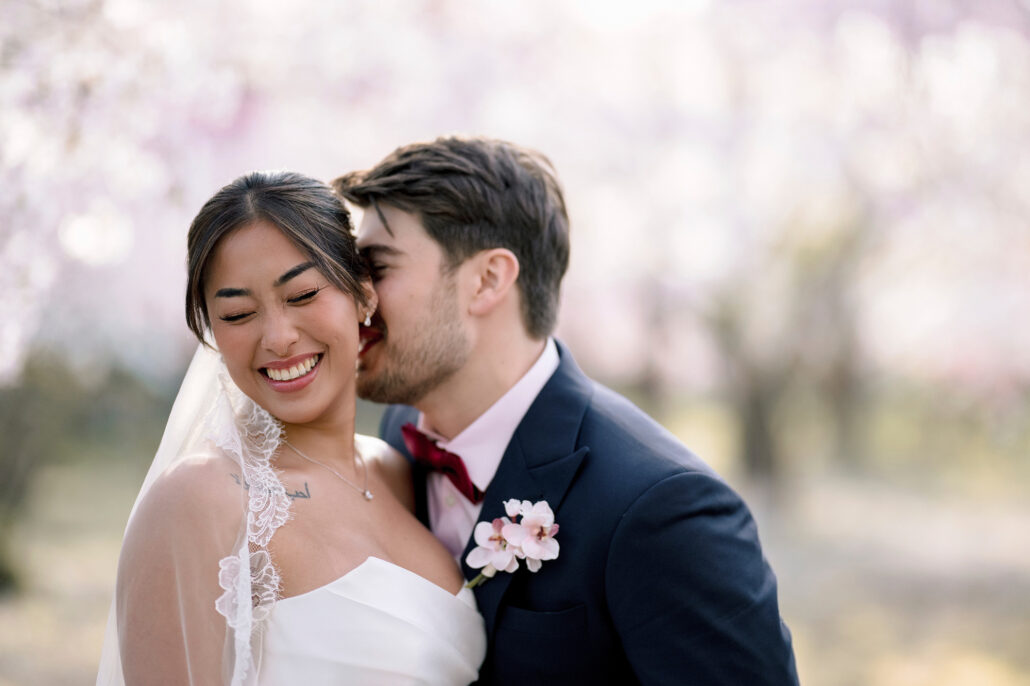 The groom kisses the smiling bride on the cheek, cherry blossom soft behind them in Aomori.