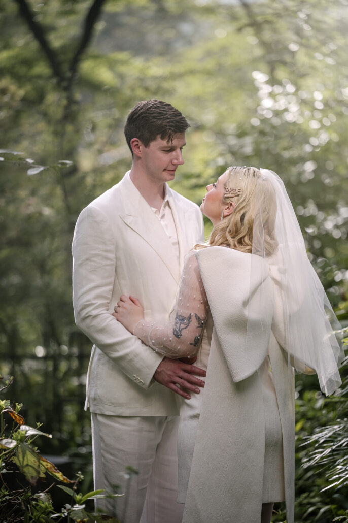The bride turns to the groom in deep summer green, the bow of her dress and her veil catching the light on an August elopement.