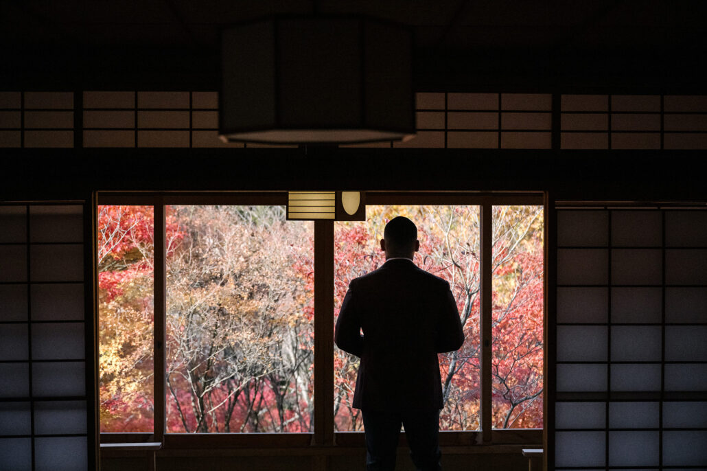 The groom stands in a dim traditional room looking out through tall windows at maple trees in full autumn colour, the season at its peak in Japan.