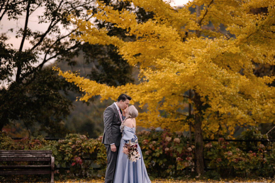 The groom kisses the bride under a blazing yellow ginkgo, her bouquet of burgundy and blush flowers, at an autumn wedding in Japan.