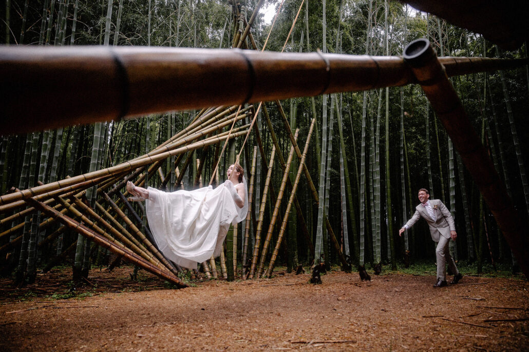 In a bamboo forest, the bride lies back on a sculpture of bamboo poles as if floating while the groom leans toward her.