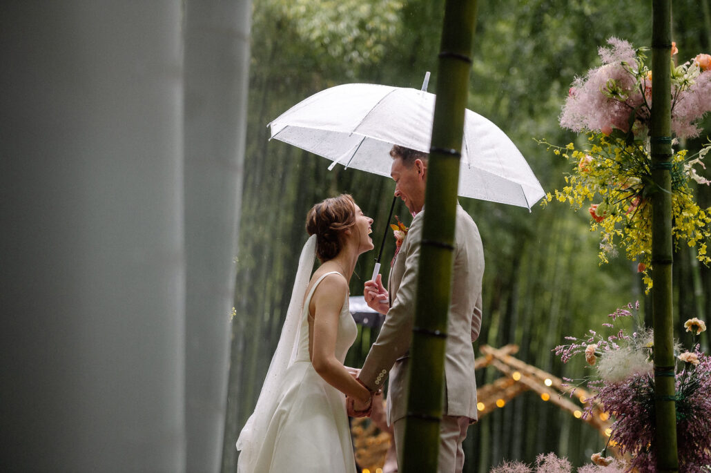 The bride looks up at the groom under a clear umbrella as rain falls on their wedding ceremony in a bamboo forest in Japan.