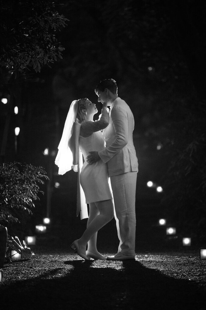 The bride reaches up to the groom's face between small lanterns on a garden path, a black and white night at Jinya.