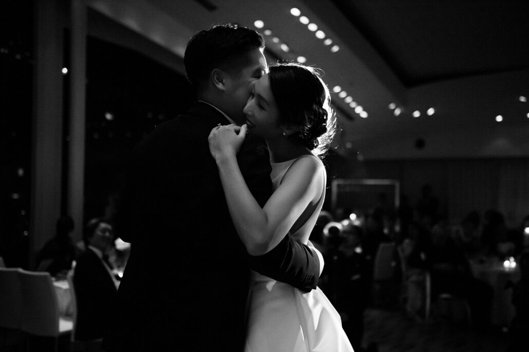 In black and white, the groom kisses the bride's cheek as she laughs behind her hand during their first dance in a darkened ballroom.