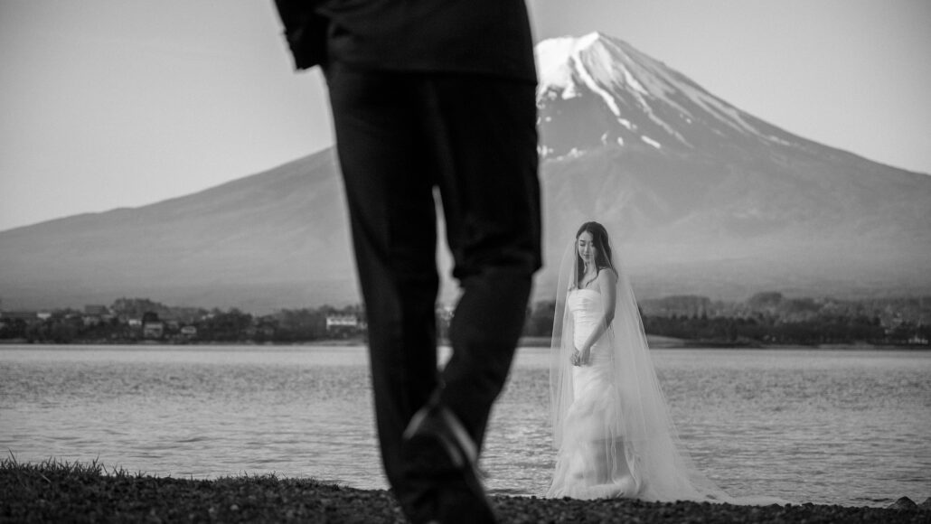 A black and white frame of the bride at the shore with Mt Fuji behind, the groom's legs striding past in the foreground.