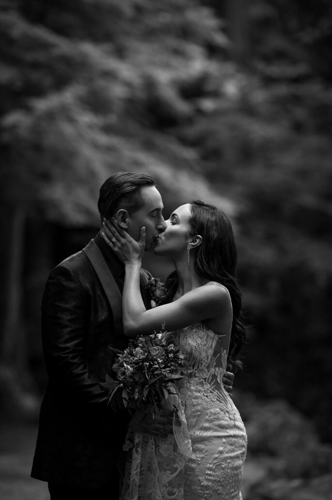 A black and white kiss in the forest, the bride's hands on the groom's face and her bouquet between them, on their wedding day in Japan.