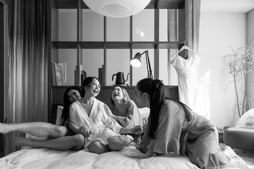 In black and white, the bride and her friends collapse laughing on the bed as they get ready, her dress hanging by the window.