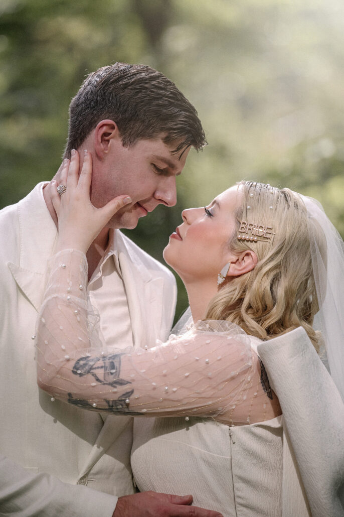 A close portrait of the bride holding the groom's face, the tattoos on her arm showing through her sheer sleeve in the garden.