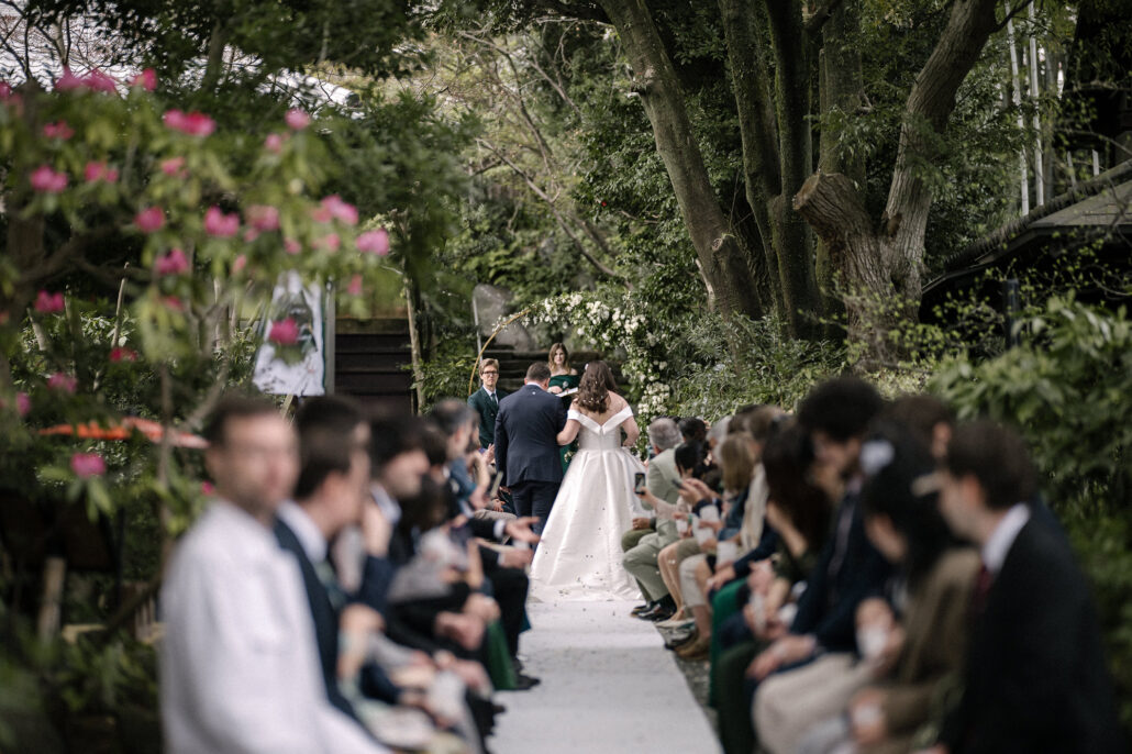 Guests in the foreground watch as the bride reaches the far end of the white aisle under tall trees, a garden wedding in Japan.