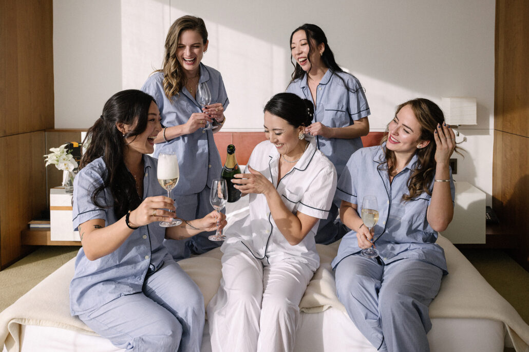 The bride and her friends laugh on the bed with champagne glasses in matching pyjamas, getting ready at Andaz Tokyo.