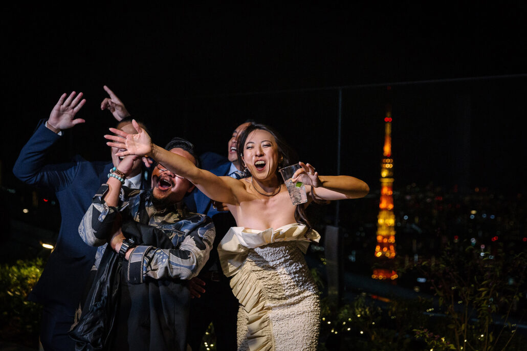The bride throws her arms out with her friends on a Tokyo rooftop at night, Tokyo Tower lit orange behind them.