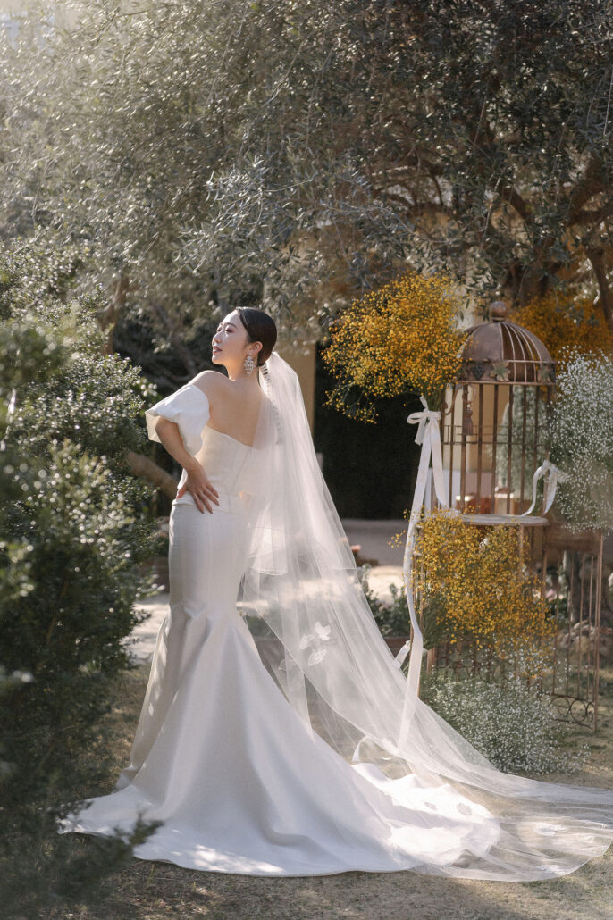 The bride laughs with her hand on her hip in a garden of olive trees and yellow mimosa, her long veil trailing behind her.