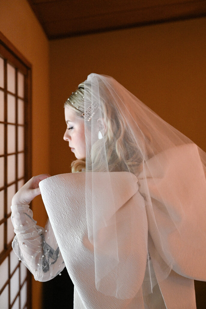 The bride getting ready in warm window light at a ryokan, her veil falling over the bow of her short wedding dress.