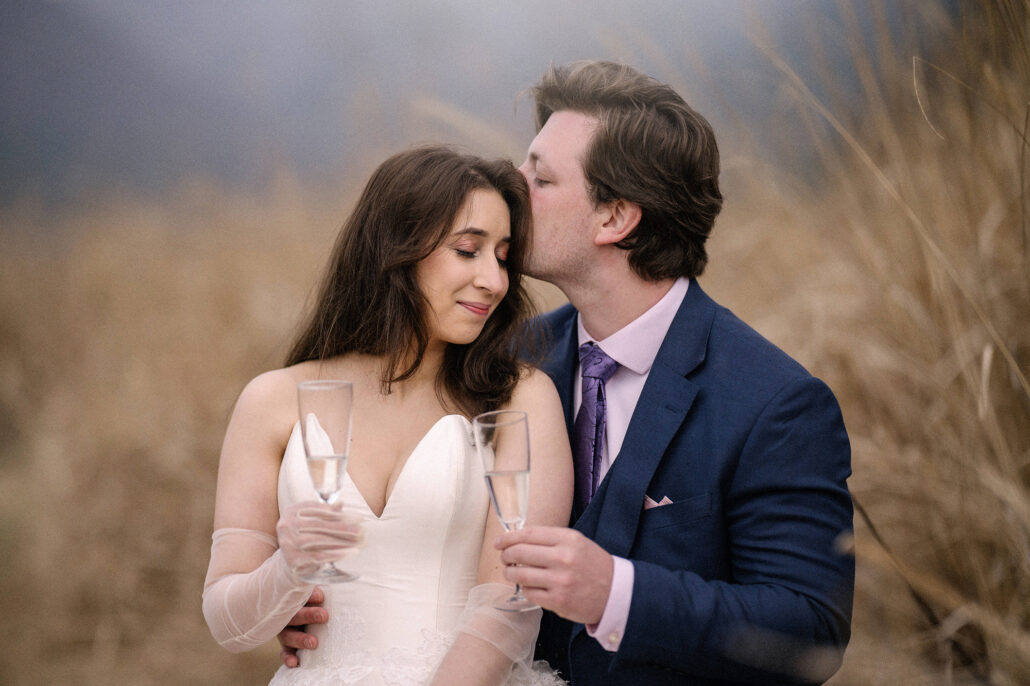 The groom kisses the bride's temple as she smiles, champagne glasses in hand, after their elopement vows at Lake Kawaguchiko.