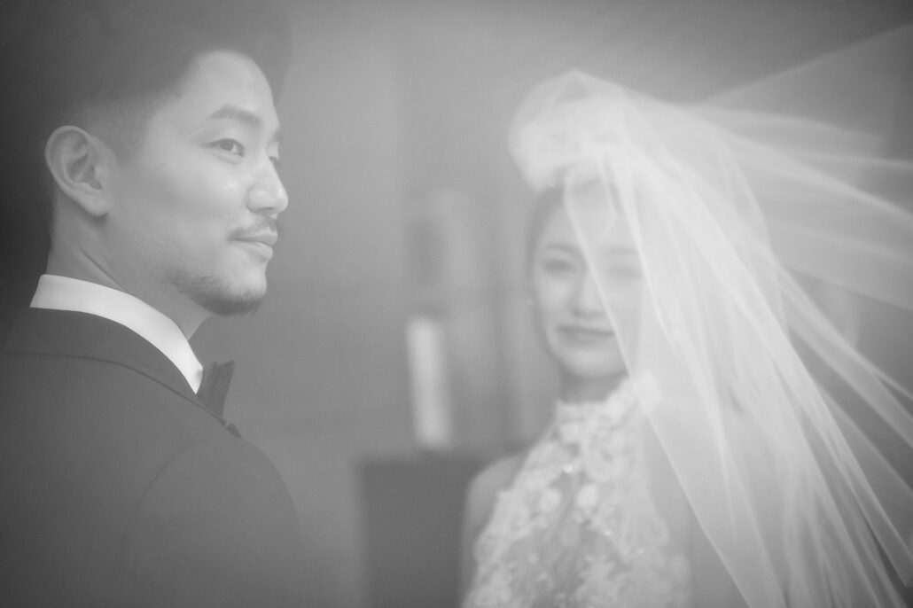 In black and white, the groom looks ahead as the bride smiles behind her veil.