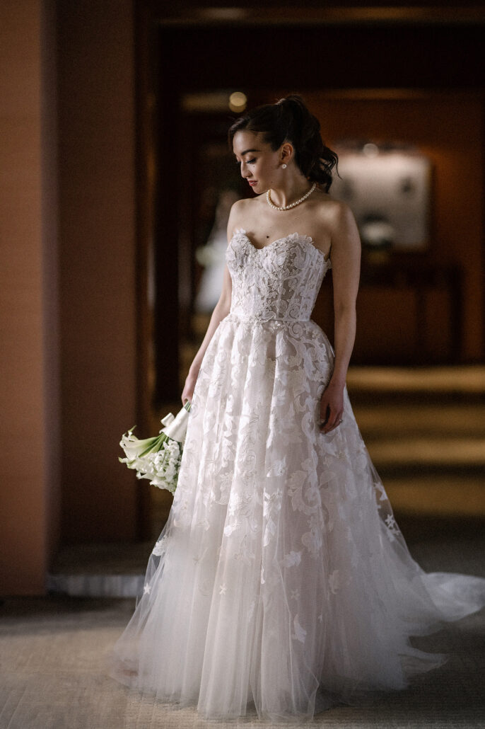 The bride stands in a warm hotel corridor in a floral tulle gown, holding white calla lilies and looking down.