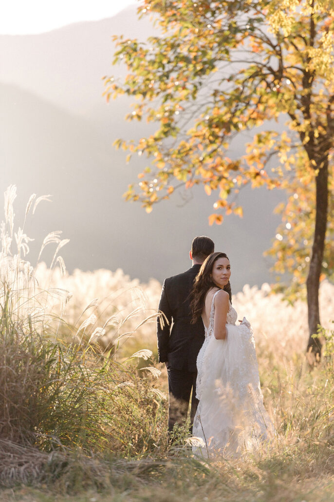 The bride looks back over her shoulder beside an autumn tree and silver pampas grass, the groom behind her, at a Lake Kawaguchiko wedding.