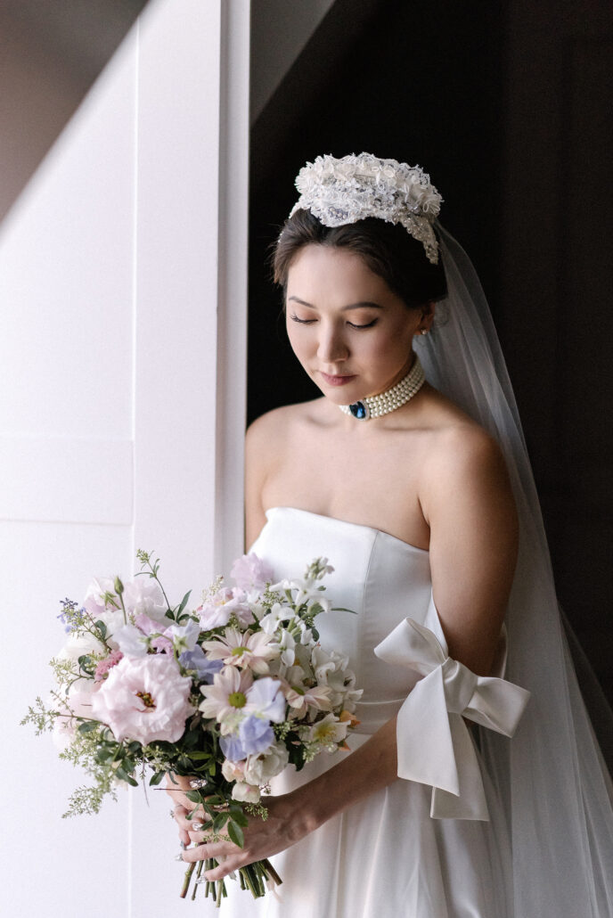 The bride looks down at her bouquet in the light of a tall doorway, a pearl choker at her throat and a lace crown in her hair.