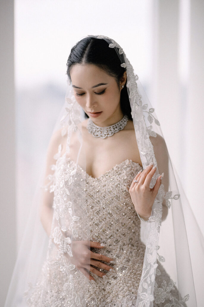 The bride stands by a tall window in a lace mantilla veil and a diamond necklace, one hand at her bodice.
