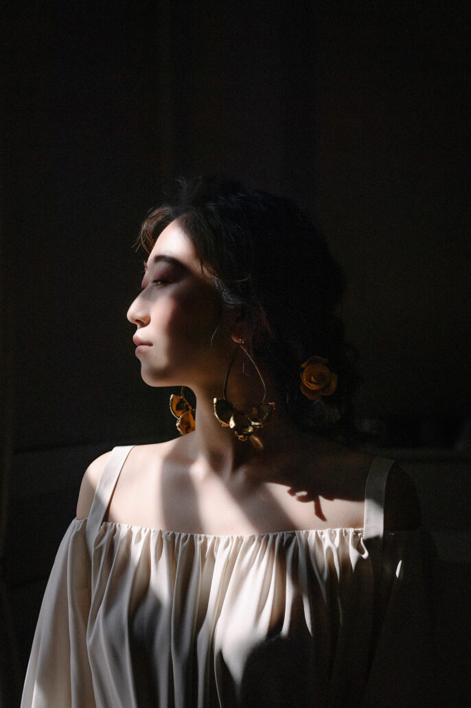 The bride turns her face to a shaft of window light, flowers pinned in her hair, the room dark around her.