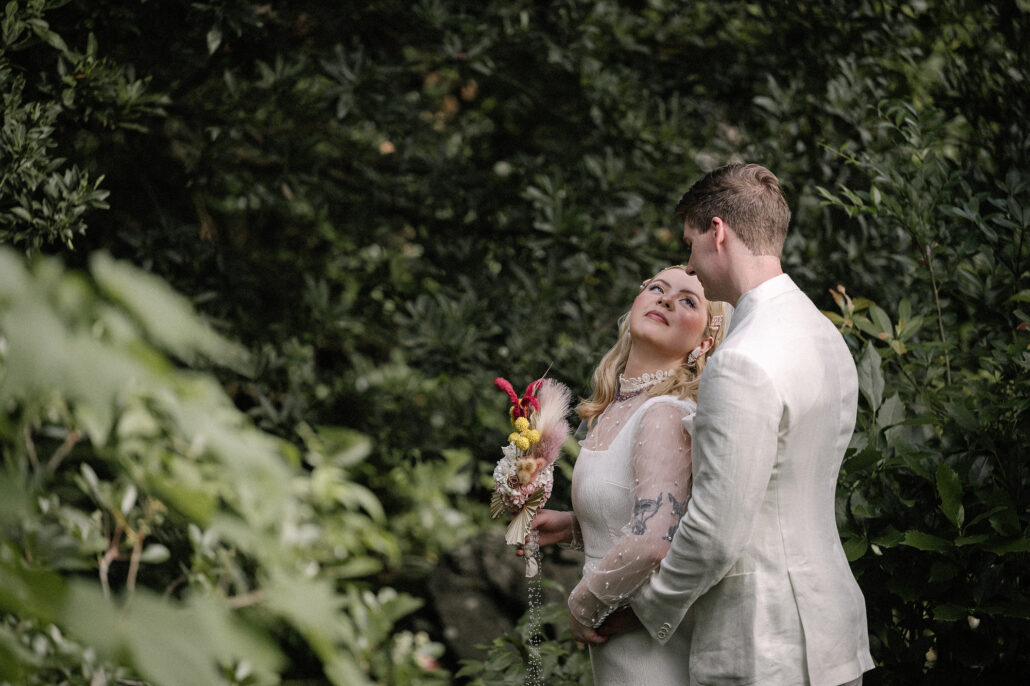 The bride leans into the groom and holds her pink, red and yellow handmade bouquet among the leaves of a Japanese garden.