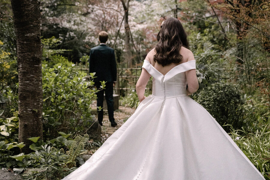 The bride walks toward the groom along a garden path under cherry blossom, her satin train spread wide, a cherry blossom wedding at Jinya.