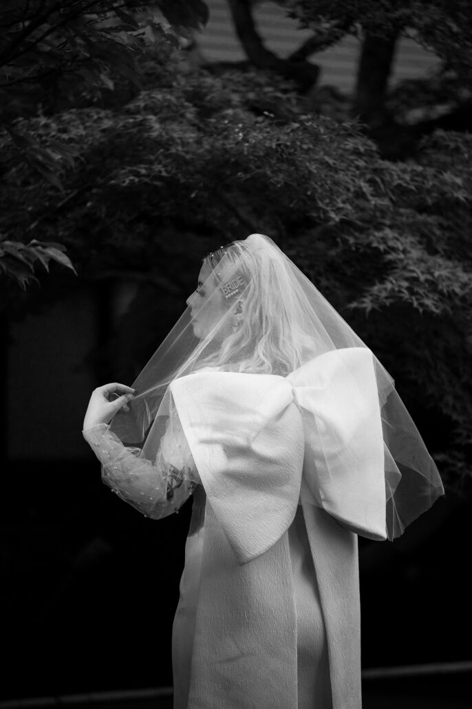 A black and white view of the bride from behind, her veil and the large bow of her dress against dark garden trees.